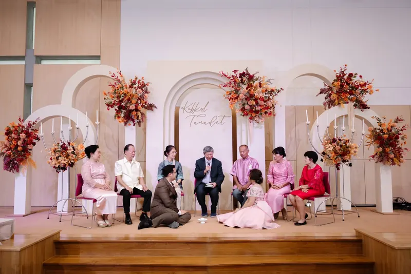 พิธีช่วงเช้าบนเวทีซุ้มโค้งกับเชิงเทียนและดอกไม้โทนส้ม ผู้ใหญ่ทั้งสองฝ่ายนั่งพร้อมหน้า