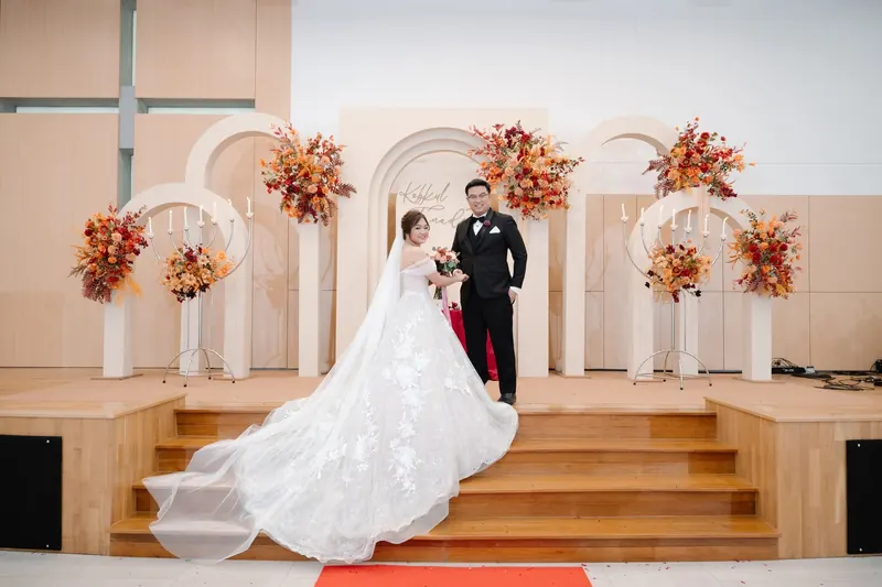 คู่บ่าวสาวบนเวทีงานเลี้ยง ซุ้มโค้งสีครีมกับเทียนและดอกไม้โทนส้ม